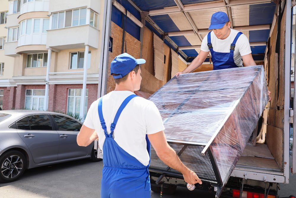 Two movers in blue uniforms and caps carefully unloading a large, plastic-wrapped piece of furniture from the back of a moving truck.