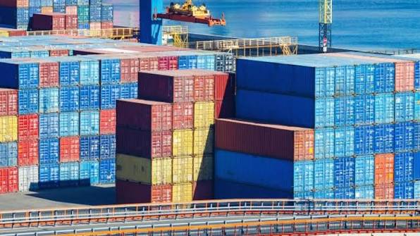 Stacks of colorful shipping containers at a port terminal with a cargo ship and blue water in the background.